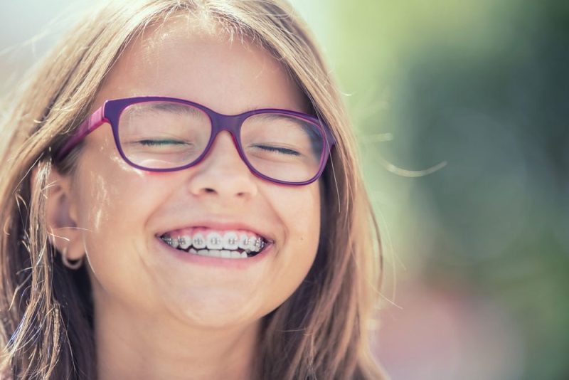 Close-up photo of a young girl with glasses laughing and showing her metal orthodontic braces
